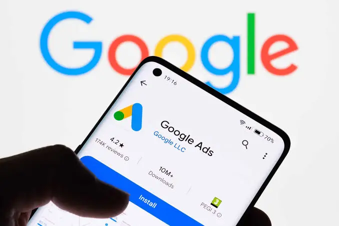 A smartphone held in a hand displaying the Google Ads application installation page in an app store against a background with the colorful Google logo.